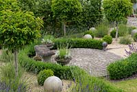 Un patio circulaire avec banc en pierre entouré de haies basses, de normes de laurier portugais et de sphères en pierre.