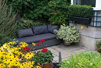 Un patio en contrebas avec canapé d'extérieur. Des fougères peintes japonaises sont plantées dans la zone ombragée derrière et des pots avec des pélargoniums brillants offrant une couleur durable.