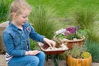 Une jeune fille avec 3 jardins miniatures en pot: bol à herbes en bois, bol alpin et brouette planté de plantes succulentes.