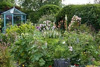 Un jardin de chalet avec parterre de fleurs mélangé coloré de Rosa 'Phyllis Bide' sur l'obélisque, Rosa 'Fantin Latour', poker rouge chaud, iris drapeau, géranium et cosmos. Derrière, une maison verte à pans de bois peints.