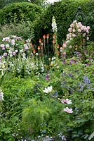 Un jardin de chalet mixte coloré avec un parterre de fleurs mélangé coloré de Rosa 'Phyllis Bide' sur l'obélisque, Rosa 'Fantin Latour', poker rouge chaud, iris drapeau, géranium et cosmos.