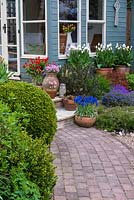Un chemin pavé menant à un pavillon d'été peint et un patio surélevé avec des pots en terre cuite plantés de Tulipa 'Coquette blanc', Muscari botroides 'Superstar' et Armeria maritima.
