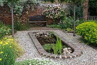 Un jardin clos avec un étang bordé de briques entouré de gravier, avec Rosa 'Phyllis Bide' formée derrière.