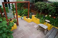 Vue d'en-haut. Sol en pierre d'aménagement paysager, murs bas peints en jaune, séparateur de jardin en bois créant deux pièces et plantation de feuillage vert.