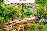 Coin repas extérieur dans un coin isolé du jardin familial avec table, chaises et parasol en bois.