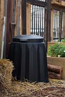 Un bac à compost en plastique noir autoportant contre un mur en bois dans un jardin communautaire.