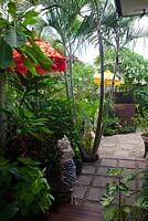 Passerelle avec des pavés de grès carrés remplis de galets de rivière dans un jardin luxuriant avec des palmiers, un Plumeria, un frangipanier, trois statues balinaises en pierre sculptée et deux parasols de temple, l'un rouge et l'autre jaune.