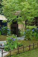 Détail d'un 'jardin d'été japonais', conçu par Saori Imoto, montrant une clôture basse en bambou au premier plan derrière laquelle se trouve une zone doucement ondulée recouverte de mousse et coupée en deux par un chemin de gravier sinueux serti de tremplins en granit, avec un fond en Acer écrans palmatum et 'bambou'. À gauche, un pavillon d'été de style japonais. Hampton Court Flower Show, juillet 2016
