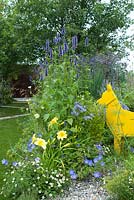 The Dog's Trust: 'It's a Dog's Life', conçu par Paul Hervey-Brookes montrant la silhouette d'un chien en acier peint en jaune reniflant les plantes. À gauche, un chemin en zigzag traverse la pelouse. Les acers multi-tiges fournissent un fond vert. Les plantes comprennent Agastach 'Black Adder', Verbena bonariensis, Agastache 'Blue Fortune' et Campanula latiflora 'Prichard's Variety' avec hémérocalles, géranium et erigeron au premier plan. Hampton Court Flower Show, juillet 2016.