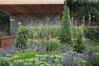 Un foyer extérieur en ardoise avec un toit en bois en porte-à-faux et un stockage de bûches de banc entouré d'herbes, d'herbes, d'arbustes et de vivaces - RHS Tatton Flower Show 2011, Cheshire