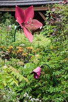 Sculptures de fleurs en acier peint dans un parterre de fleurs près de la maison de style chalet entourée de ligularia orange, eupatoriums et bambou. Hunting Brook Garden, Co Wicklow, Irlande