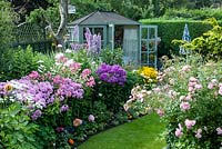 Jardin arrière coloré avec des parterres de fleurs mélangés remplis de plantes annuelles, d'arbustes et de vivaces, dont Rosa 'The Fairy', Delphinium 'Sweet Sensation', Phlox 'Elizabeth'