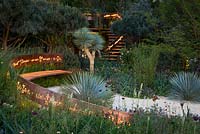 Le jardin Winton Beauty of Mathematics éclairé la nuit, RHS Chelsea Flower Show 2016. Symboles mathématiques illuminés coupés en bande de cuivre traversant le jardin formant l'arrière du banc et la rampe pour l'escalier. Pinus sylvestris 'Glauca' - pin sylvestre bleu, Yucca rostrata - Yucca à bec
