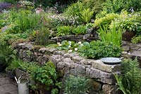 Un jardin de chalet avec étang surélevé, jardin en pot cultivant des fruits, des herbes et des légumes et un grand parterre de fleurs en pente avec des vivaces mixtes.