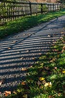 Chemin goudronné avec des ombres de la clôture et des feuilles tombées sur une journée d'automne ensoleillée.