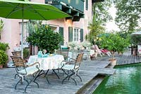 Sur la terrasse en bois entre une maison et un étang de baignade, des meubles de jardin en bois, un muret en pierre et des plantes en pots, dont des agrumes