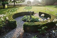 Parterre de fleurs rond avec roses, haie de buis et cadran solaire central. Jardin Yvan et Gert en Belgique.