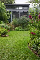 Vue depuis l'arrière du jardin vers l'arrière de la maison, montre des parterres de jardin incurvés, une pelouse de buffle à feuilles souples et une variété de plantes herbacées à fleurs.