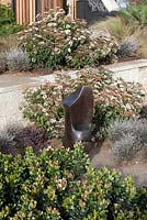 Une sculpture en bronze avec une plantation mixte d'arbustes et de plantes basses représentant Rhaphiolepis 'Snow Maiden', Indian Hawthorn, Virburnum cultivar et Corokia cotoneaster