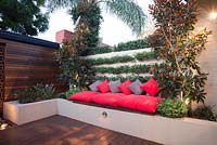 Cour intérieure avec banquette de jardin surélevée avec coussins gris et rouges. Un mur de briques avec Trachelospermum jasminoides et Star jasmine