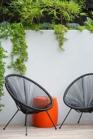 Une terrasse en bois avec des chaises Acapulco noires, une table de tambour en plastique orange devant un ciment peint en gris a rendu un mur de soutènement avec des plantes succulentes débordant sur le mur et un écran de tisserands élancés en bambou.