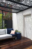 Terrasse en bois avec salon extérieur avec coussins et pergola avec toit en métal à motifs géométriques découpés au laser projetant des ombres graphiques sur un mur.