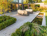 Un patio, avec des sièges et une rigole, dans un jardin de campagne moderne du Cheshire, conçu par Louise Harrison-Holland. La plantation comprend Amelanchier lamarckii, la couverture de boîte et Stipa tenuissima