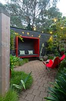 Zone pavée avec un mur en bois peint en noir avec une cosse assise rouge attachée à une chaise papillon rétro en toile rouge et une collection de grands pots avec un érable et diverses plantes succulentes.