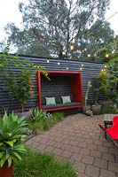 Zone pavée avec un mur en bois peint en noir avec une cosse assise rouge attachée et une collection de grands pots avec un érable et diverses plantes succulentes. Un parterre de fleurs d'herbe indigène australienne, Lomandra 'Tanika '.