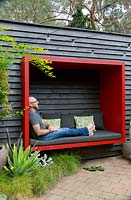 Le propriétaire et concepteur du jardin, Steven Wells, se détend dans une nacelle en bois peint en rouge qu'il a construit, attachée à un mur en bois peint en noir.