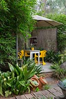 Terrasse en bois recouverte d'un grand parapluie blanc avec des chaises rétro en plastique jaune vif et une table en bois, recouverte de stores allumettes en bambou et encadrée par une touffe de bambou et Aspidistra elatior, usine de fonte.