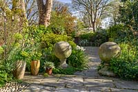 Une vue sur le jardin avec Euphorbia, le lierre envahissant le mur de pierre, des arbres matures, des plantes en pot et deux globes de pierre architecturale sur des piédestaux