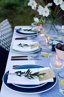 Table à manger d'extérieur habillée dans des tons de bleu et avec un vase d'anémones japonaises. Des poches sont faites dans les serviettes pliées pour y mettre la carte de visite et chaque décor est fini avec une branche d'olivier. Des bougies chauffe-plat dans des verres éclairent la table au crépuscule