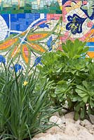 The Viking Cruises Garden of Inspiration - Un mur de mosaïque incurvé inspiré par les caractéristiques de Gaudi dans le jardin Vicking Cruise avec Aeoniums, Senecio mandraliscae marine poussant dans le gravier et un oranger,