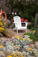 Jardin avant sur le thème de la mer avec chaise Adirondack blanche, plantée de plantes côtières, dont Cineraria 'Silver Dust', Stipa tenuissima et Tamarisk tetandra fleurissant derrière et décorées de bouées de sauvetage et de flotteurs de pêche.