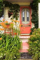 Façade de maison de style chalet ancien avec porte rouge et véranda avec hélice Hedera grimpante. Parterre de fleurs avec des arbustes Hemerocallis et Spiraea.