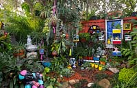 Zone arrière surélevée du jardin avec une collection éclectique d'ornements de jardin, de pots, de statues et de paniers suspendus parmi des plantes aimant l'ombre.