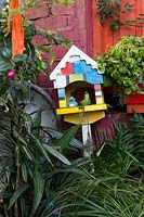 Une maison d'oiseau ornementale d'inspiration mondrienne parmi des plantes à feuilles lanières appuyées contre un mur rose et orange.