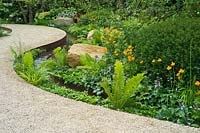 Un jardin avec chemin de gravier sinueux à côté d'un ruisseau. Les plantes comprennent Trollius, Hosta elegans, Matteuccia struthiopteris