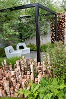 Le jardin naturellement à la mode conçu par NDG. Coin salon avec sièges en maille métallique, pergola noire, rondins de bouleau