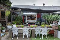Vue d'un café en plein air, avec un bar flottant pour chevaux converti et une longue table en bois avec jardinières intégrées et chaises en fonte blanche, Australie.