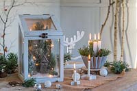 Arrangement festif utilisant du feuillage et des pommes de pin, des lumières LED, des bougies et des boules dans un cadre rustique.