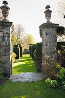 Un chemin herbeux entre des piliers en pierre surmontés d'urnes mène entre des ifs coupés vers un belvédère encadré par une paire de tilleuls blanchis. Plas Brondanw, Penrhyndeudraeth, Gwynedd, Pays de Galles
