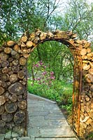 Chemin menant à travers l'arche en rondins dans le verger avec un Magnolia à fleurs roses 'Susan '. Summerdale Garden, Cumbria, Royaume-Uni.