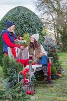 Femme donnant un panier avec des cadeaux à un ami assis sur un banc, entouré de verdure et de décorations de Noël