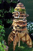Fusée multicouche en bois avec différents matériaux pour le motel des insectes, y compris des pommes de pin, des carreaux cassés, de la mousse et de l'herbe séchée. Jardin 'My Space in Space', par Stanley Road Primary School, RHS Malvern Spring Festival, Royaume-Uni.