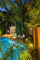 Jardin éclairé la nuit avec vue sur la piscine vers le coin salon sur terrasse en bois, clôtures et plams