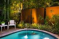 Jardin contemporain offrant intimité grâce aux murs et à la plantation tropicale. Conçu pour être utilisé la nuit avec éclairage, coin salon, terrasse en bois et piscine.
