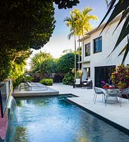 Piscine avec cascade près de la maison avec des zones pavées pour les sièges et les palmiers et la plantation tropicale au-delà