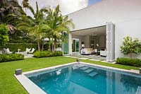 Vue sur la piscine à l'arrière de la maison avec ses portes pliantes ouvertes, au-delà des parterres de palmiers est un coin salon extérieur